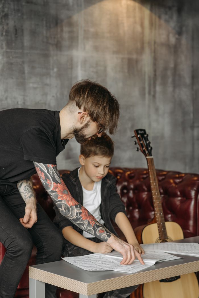 about-img A teacher guides a child through guitar sheet music in an indoor setting.